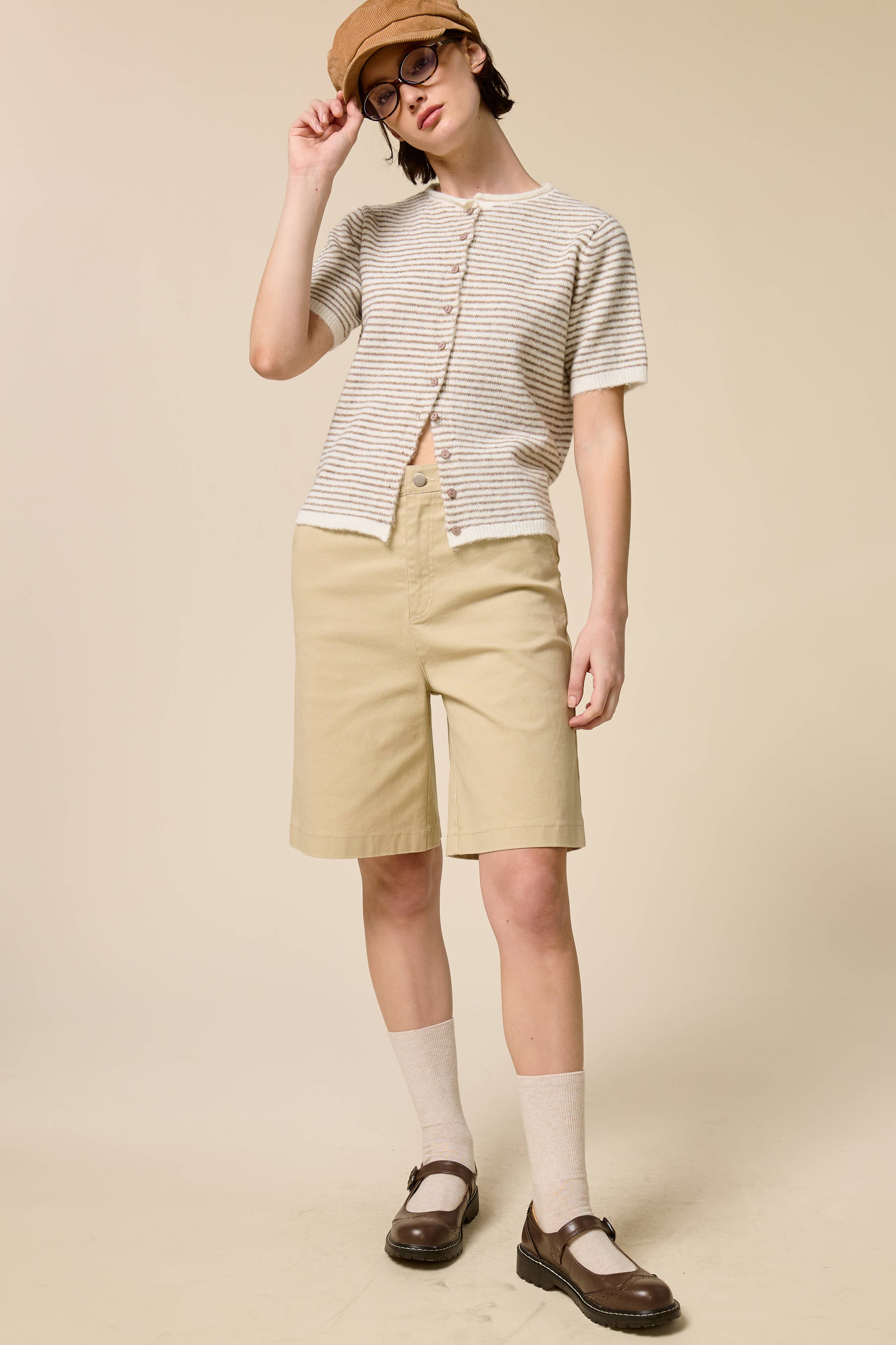 Person wearing a striped shirt, beige shorts, and a hat on a beige background