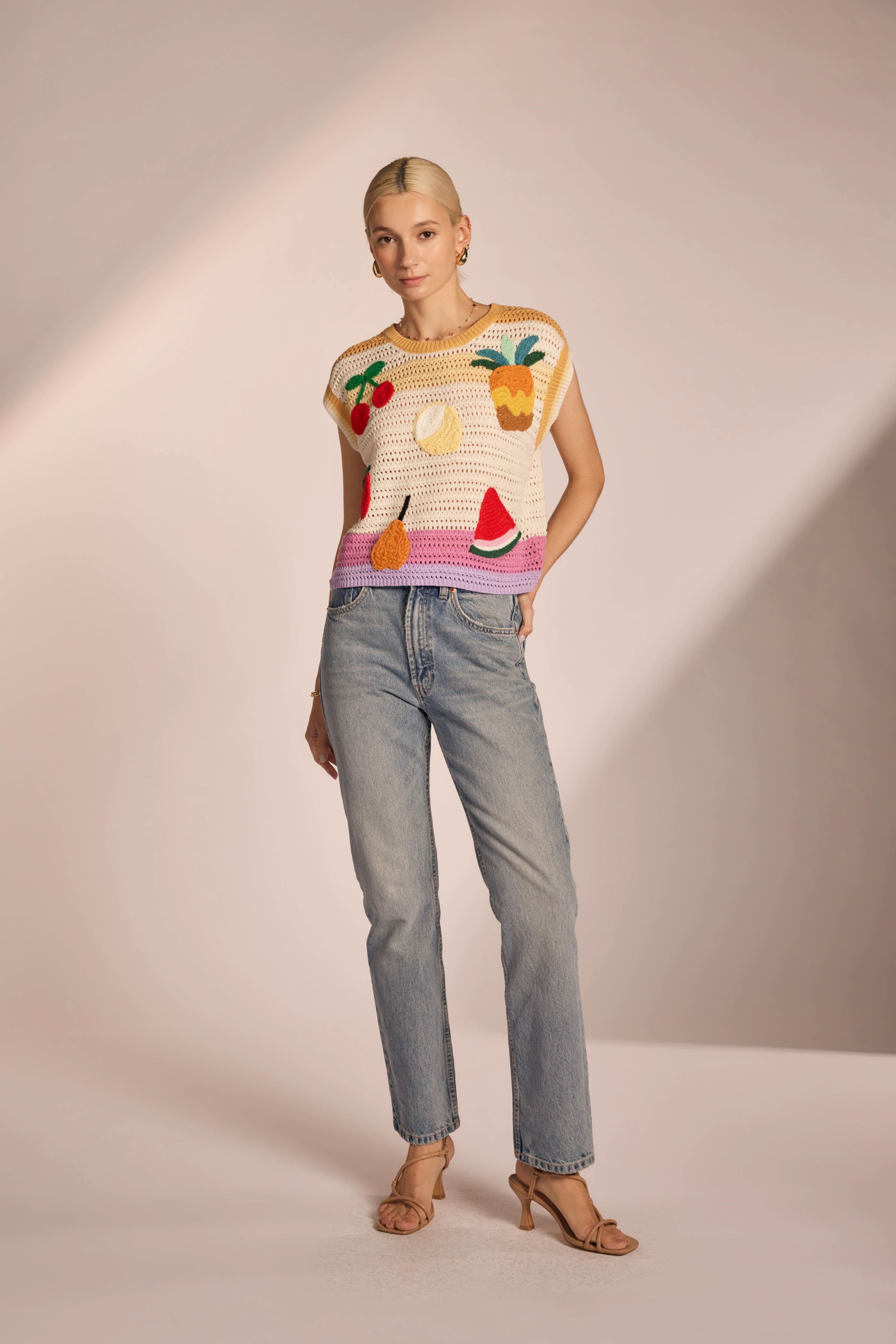 Woman wearing sleeveless, ivory, colorful, crochet, top with cherries, lemon, strawberry, watermelon, pear, and pineapple with pink, purple, and yellow horizontal stripes 