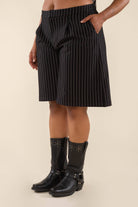 Woman wearing black vertical pin striped knee length shorts with pockets and black boots behind a blank background. 