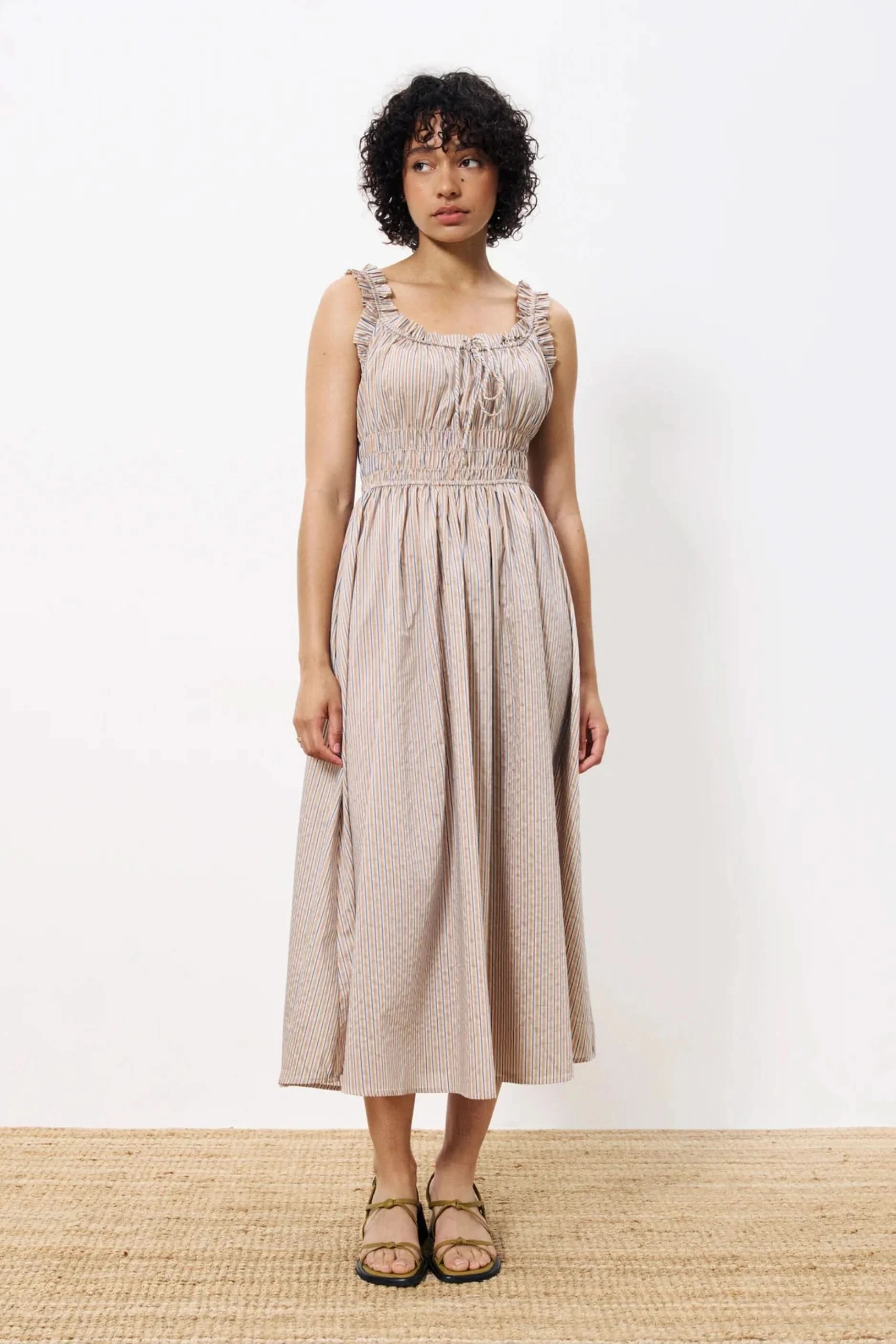 Woman wearing long flowy sleeveless dress, smocked at the waist, pinstriped in tan, royal blue and white