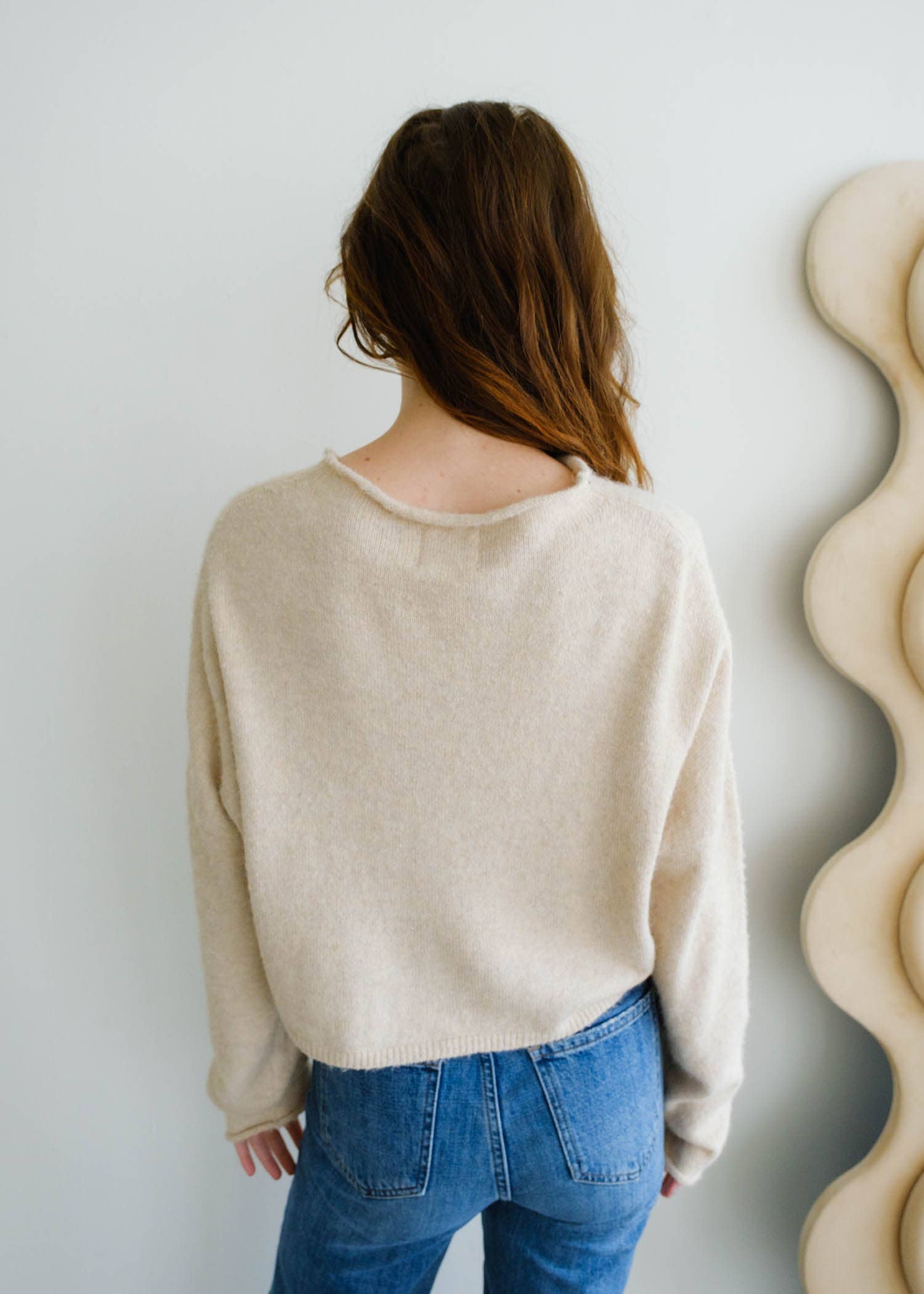 Woman facing away from the camera wearing blue jeans, and a natural beige button up piper cardigan 