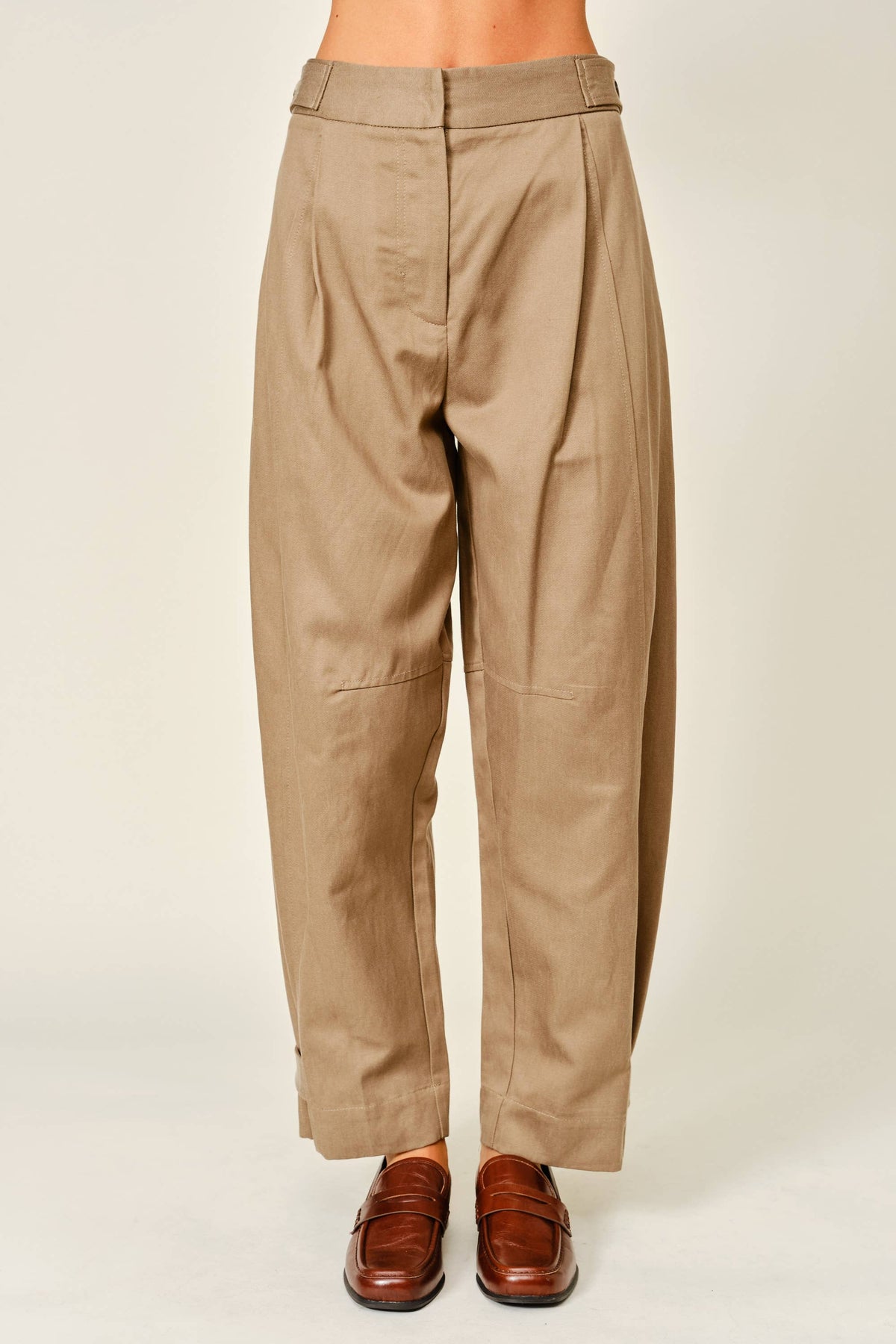 A person shown from the waist down wearing loose, high-waisted tan pants with front pleats and brown leather loafers, photographed against a plain light beige studio background.