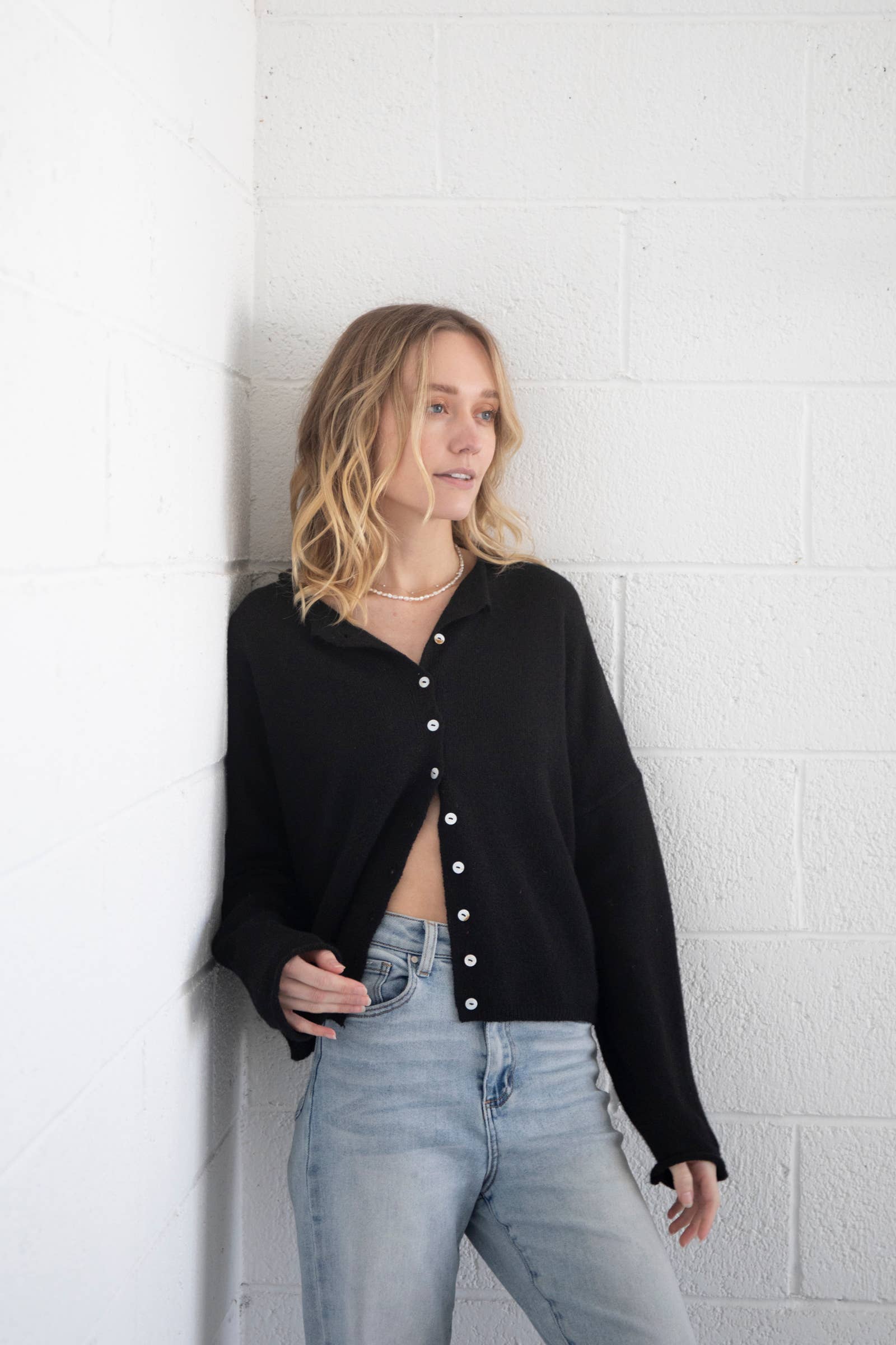 Woman wearing black cardigan with small white buttons unbuttoned halfway down and blue jeans 