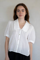 Woman standing against white background wearing white collared button down blouse with extended sleeves and vertical line details 