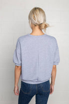 Woman facing away from the camera wearing natural grey, short sleeve, puff sleeve, blouse t-shirt top, with vertical indent line down the back 