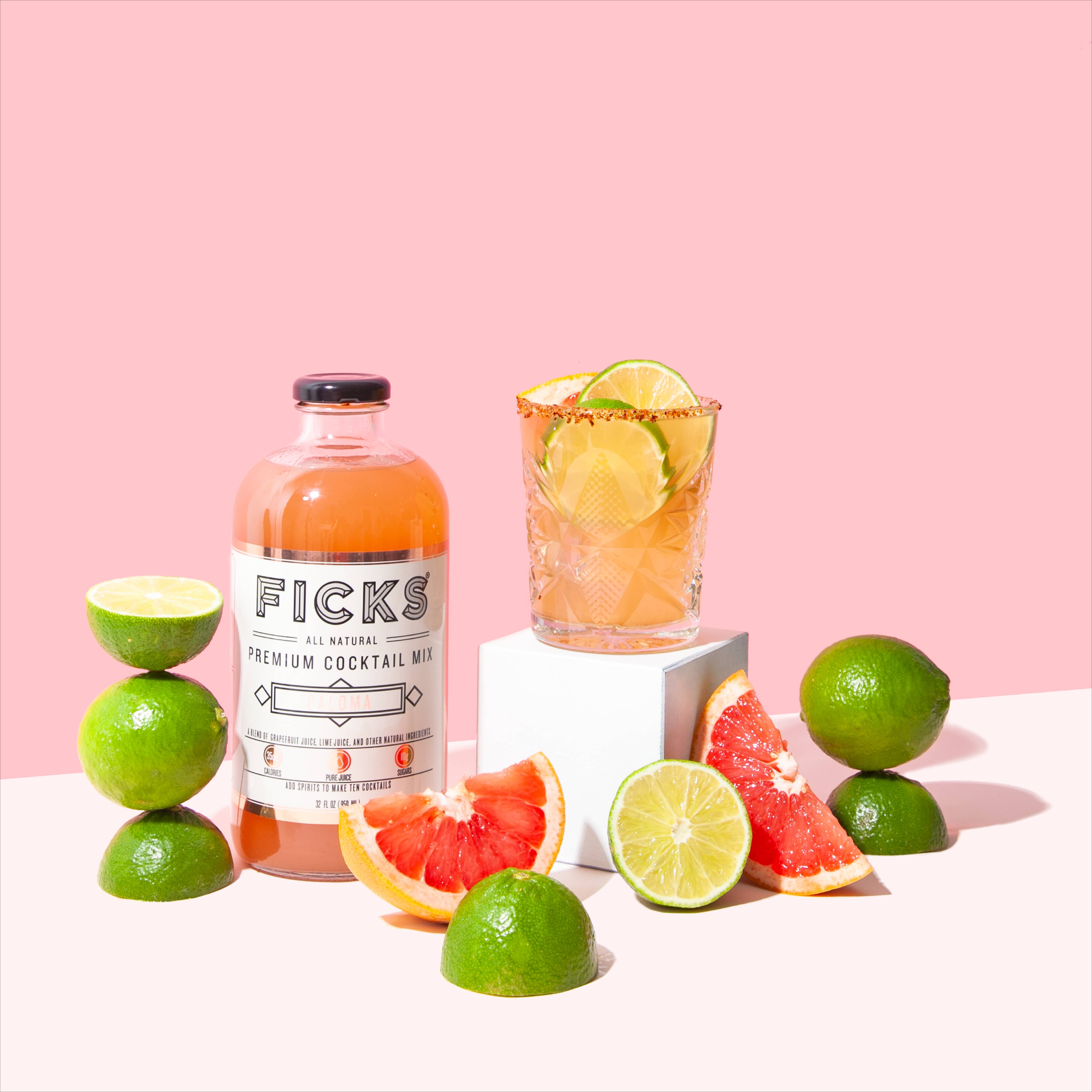 Glass bottle containing pink cocktail next to glass with cocktail ingredients. Limes and grapefruit slices for display  