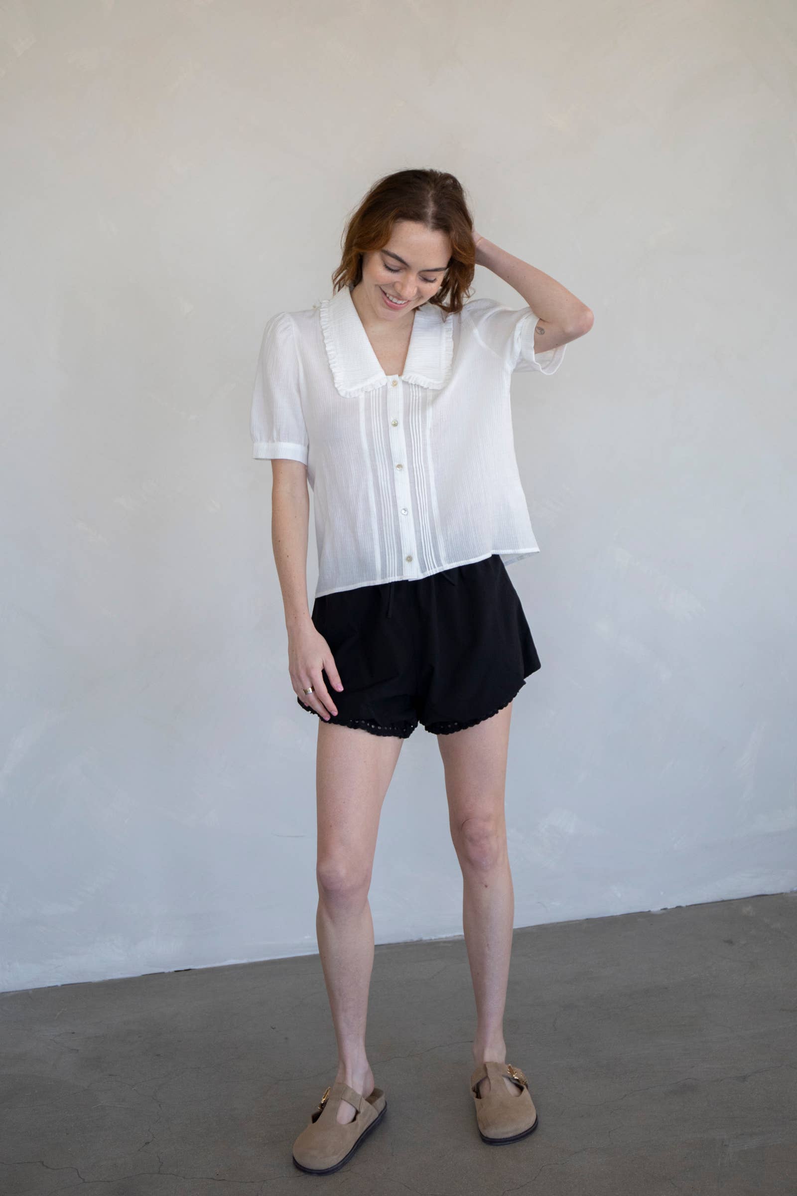 Woman standing against white background wearing tan clogs, black short, and a white collared button down blouse with extended sleeves and vertical line details 