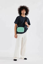 Model standing against white background wearing Green gingham BAGGU crossbody camera bag with black adjustable strap