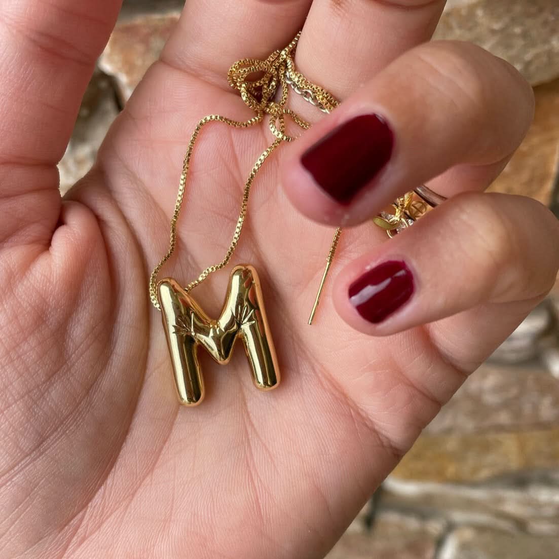 Gold necklace with a 'M' pendant held in a hand with red nail polish.