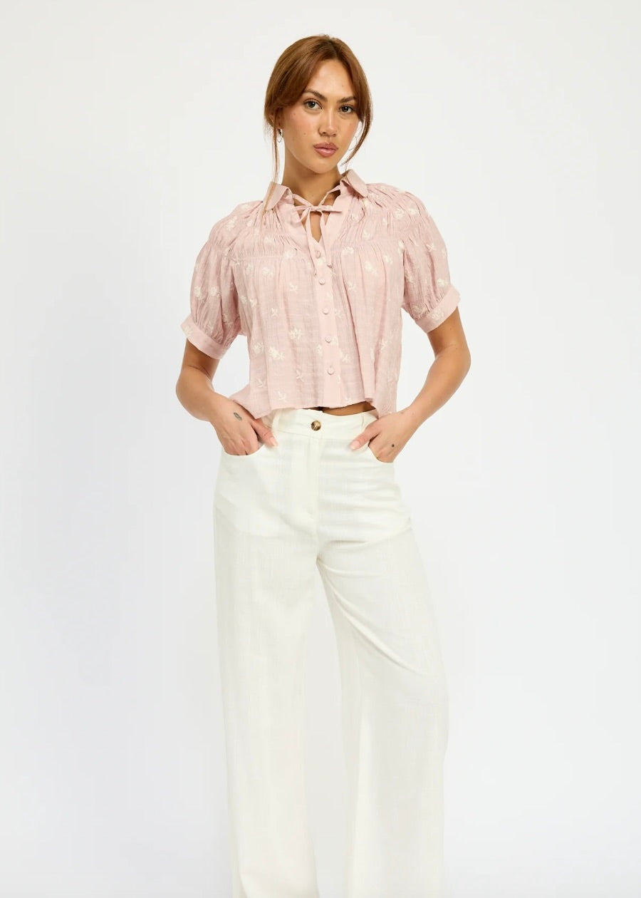 Model standing against a white background wearing white jeans and a pink flowy blouse with tulip embroidery details