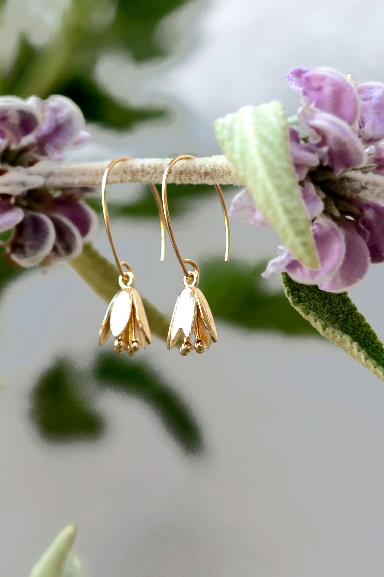 Gold flower-shaped earrings hanging on a branch with purple flowers.