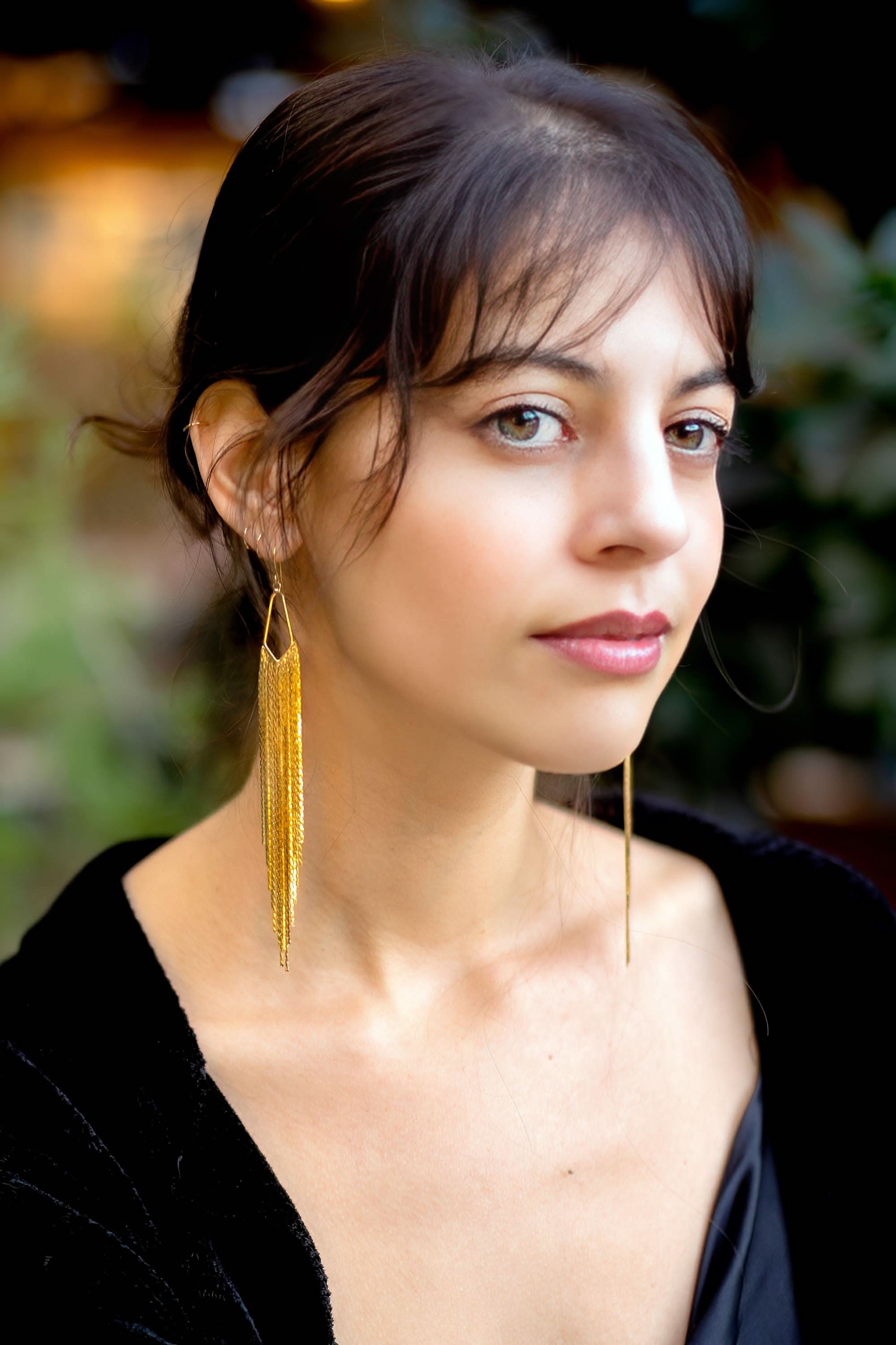 Woman wearing long gold earrings with a blurred outdoor background