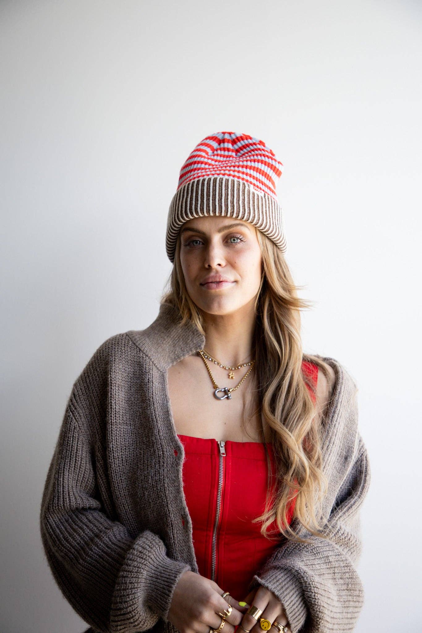 Woman wearing a red and white striped beanie, gray cardigan, and red top against a plain background