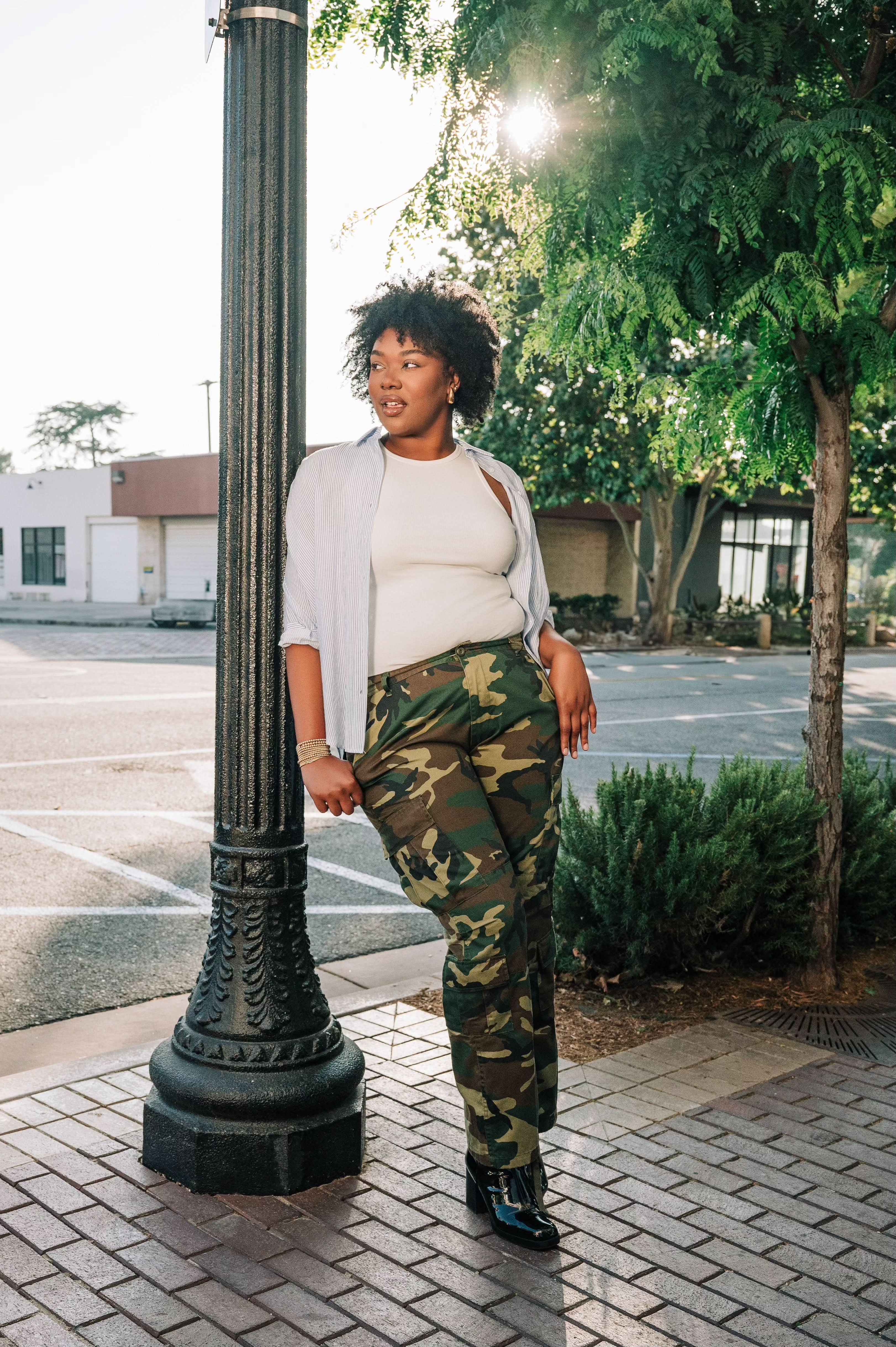 Woman wearing a white ribbed high neck tank top with a white and blue vertical button down with camo pants and black shoes. 
