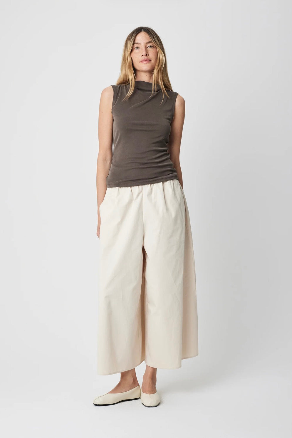 A woman modeling a sleeveless dark brown mock-neck top and cream wide-leg cropped pants with cream flats, shot against a light gray studio background.