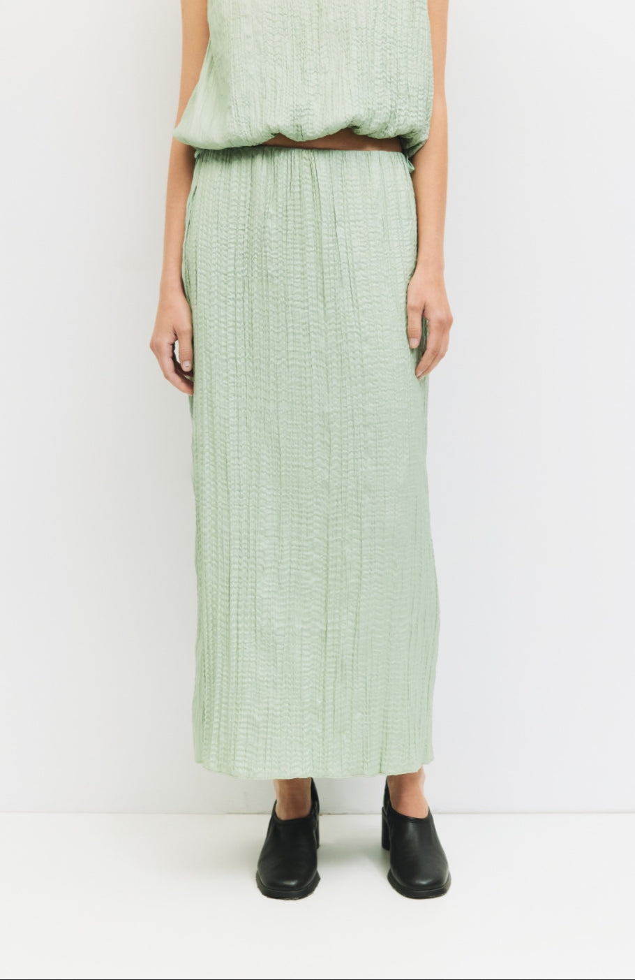 Woman standing against a white wall wearing matching sea foam, Sage green textured skirt and blouse