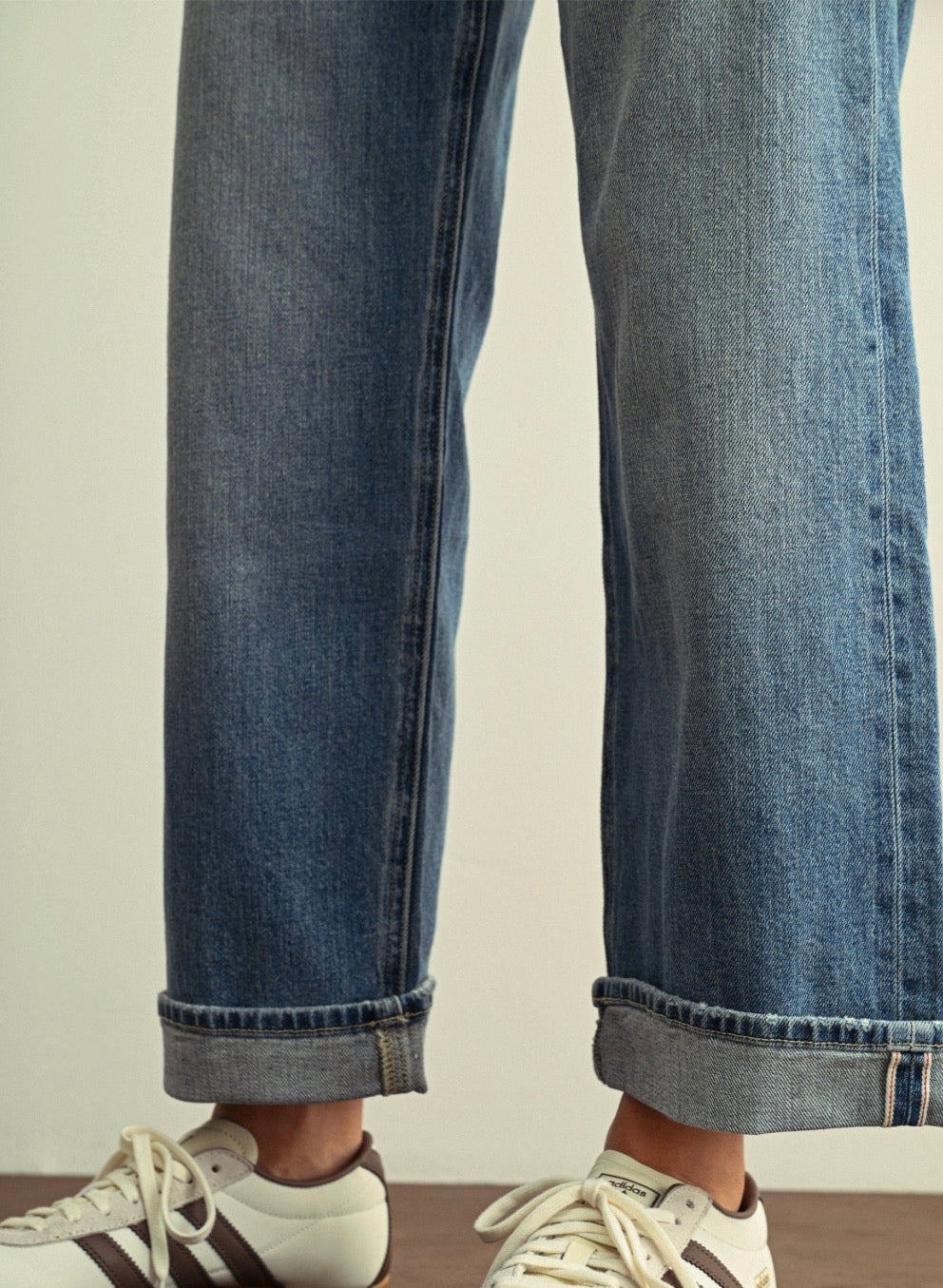 Close-up of a person wearing blue denim jeans with rolled cuffs and white sneakers against a plain light background.