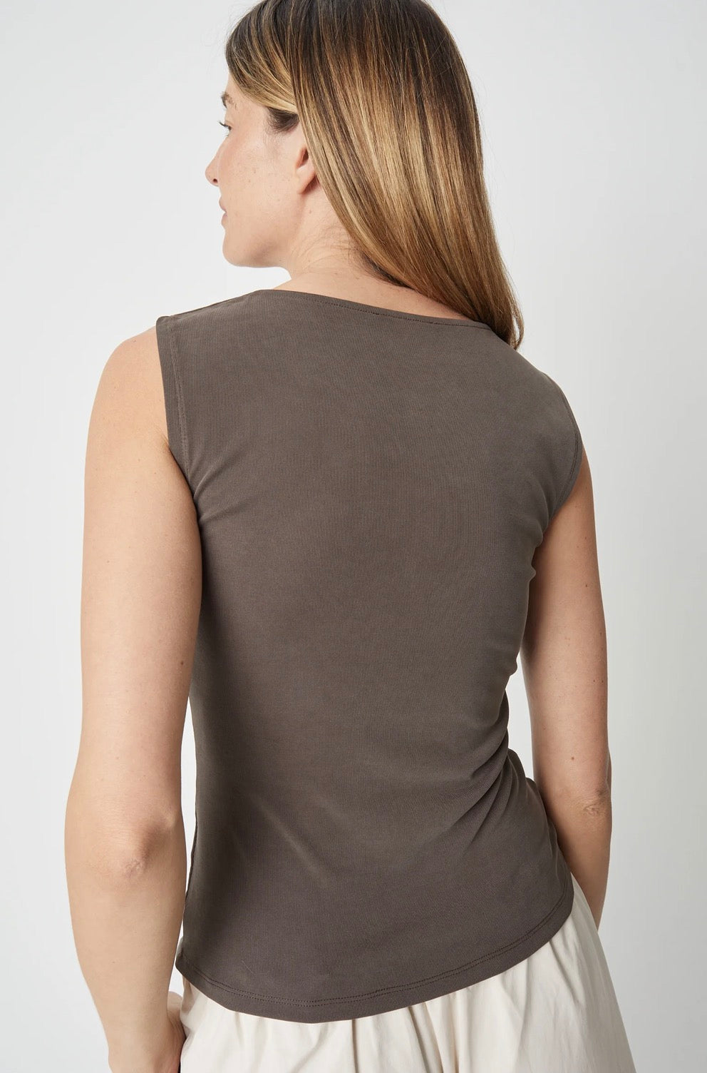 A woman shown from the back wearing a sleeveless, scoop-neck top in a muted taupe-brown color, paired with light-colored bottoms, against a plain light gray studio background.