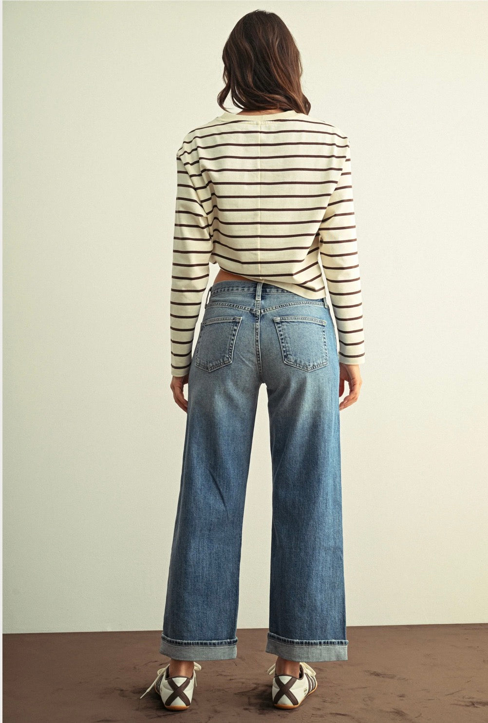 A woman viewed from behind wearing a cream and black striped long-sleeve crop top, wide-leg blue jeans, and white lace-up shoes, standing against a plain light beige wall.