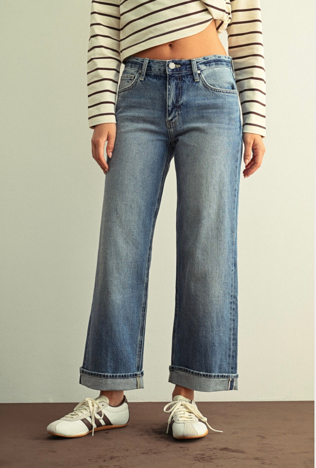 A person wearing light-wash blue wide-leg cropped jeans with rolled cuffs, paired with a cream and brown striped long-sleeve crop top and white sneakers, standing against a plain light wall.