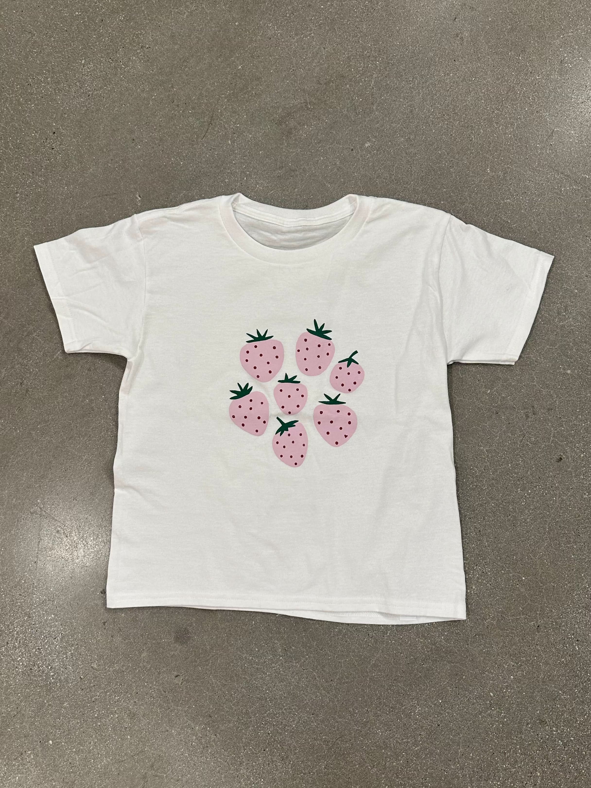 White tee-shirt with six pink strawberries on the front in front of a blank background. 