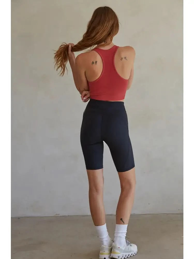 Woman wearing a red knit seamless ribbed scoop with a U-V neckline tank top with black biker shorts standing in front of a blank background. 