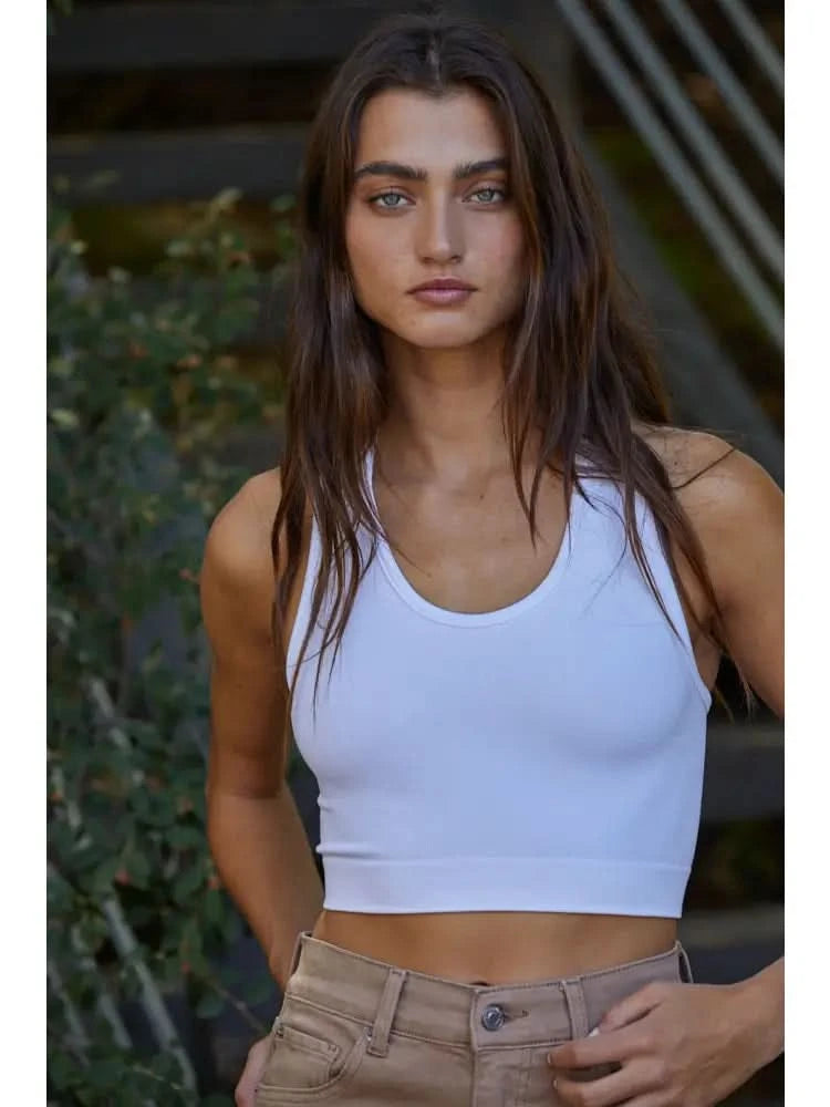 Woman wearing a white knit seamless ribbed scoop with a U-V neckline tank top with brown jeans standing behind green foliage. 