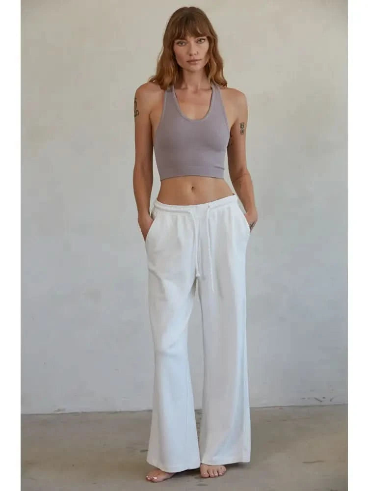 Woman wearing a mauve knit seamless ribbed scoop with a U-V neckline tank top with white drawstring sweat pants standing in front of a blank background. 