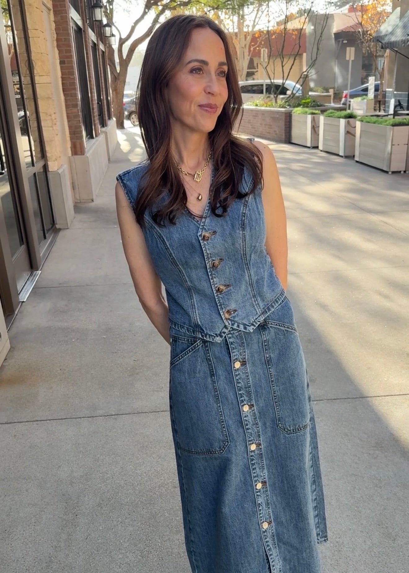 Woman smiling, standing outside wearing blue jean button up vest and matching jean skirt