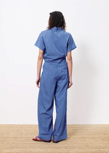 Woman facing away from viewer wearing blue botton up blouse with vertical red stripes and a front pocket. Wearing pants with the same pattern.