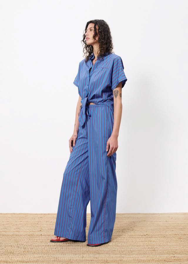 Woman wearing blue botton up blouse with vertical red stripes and a front pocket. Wearing pants with the same pattern.