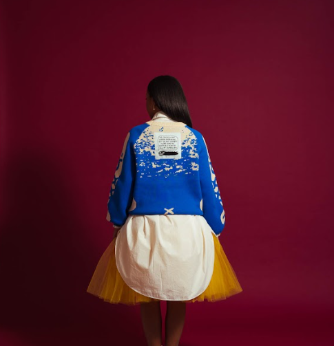 Woman facing backward wearing royal blue and white knit sweater with white oversized dress shirt underneath paired with a mustard yellow tulle skirt 
