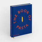 Blue hand over cookbook titled in red “THE BOOK OF PASTA” with a cut out image of a single penne noodle 