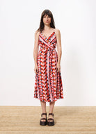 Woman wearing colorful wrap around maxi dress with tank top detail standing against a white wall on a brown  carpet. 
