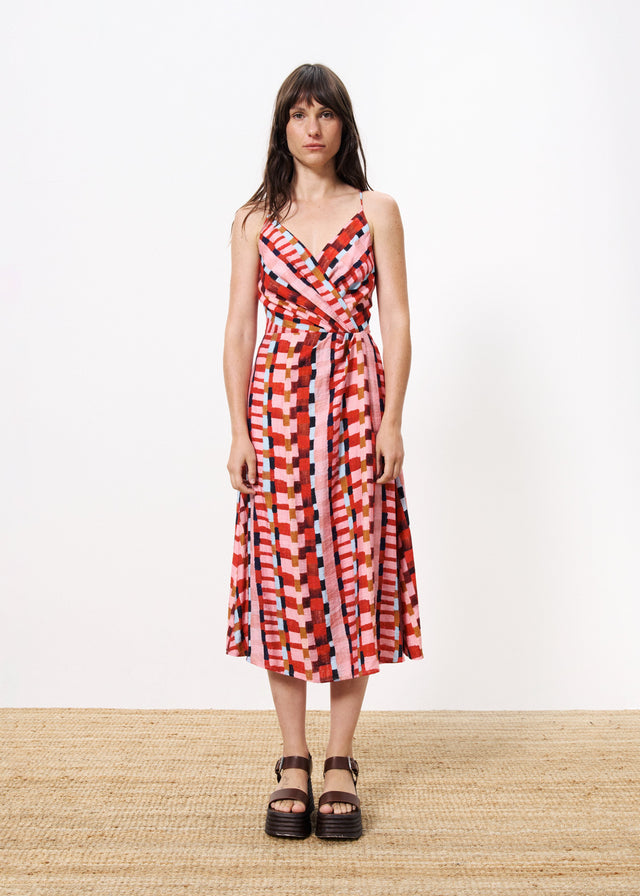 Woman wearing colorful wrap around maxi dress with tank top detail standing against a white wall on a brown  carpet. 