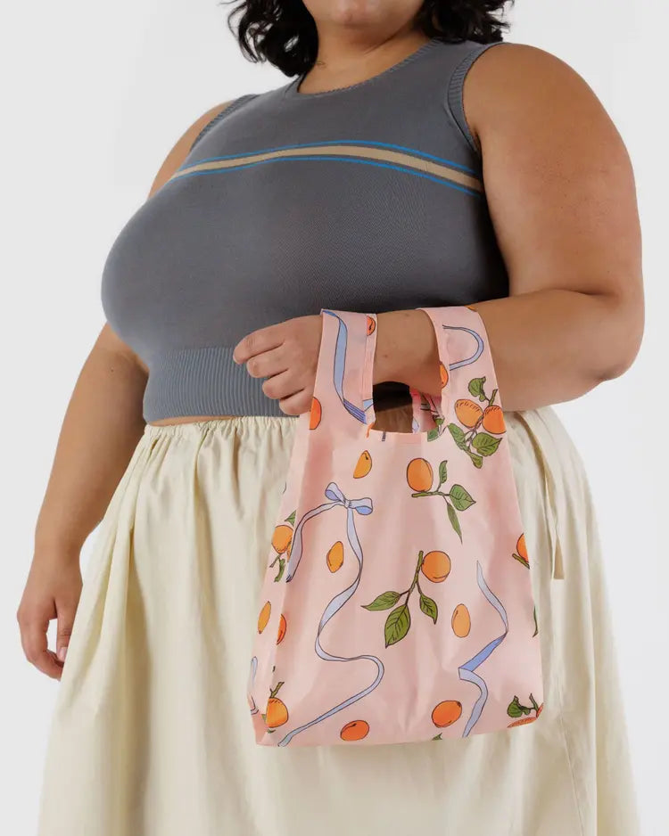 Woman wearing a grey tank top and yellow skirt holding a light pink small reuseable bag with orange apricots and blue ribbons behind a white background. 