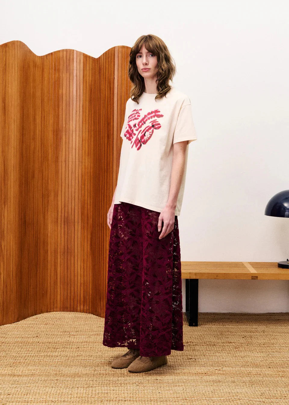 Woman wearing a white t-shirt with red text and a dark red patterned skirt standing in a room with wooden paneling.