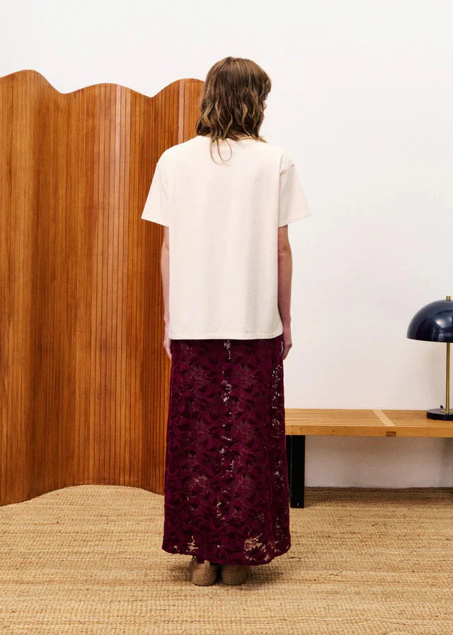 Person wearing a white top and burgundy lace skirt standing in a room with wooden paneling and a lamp.