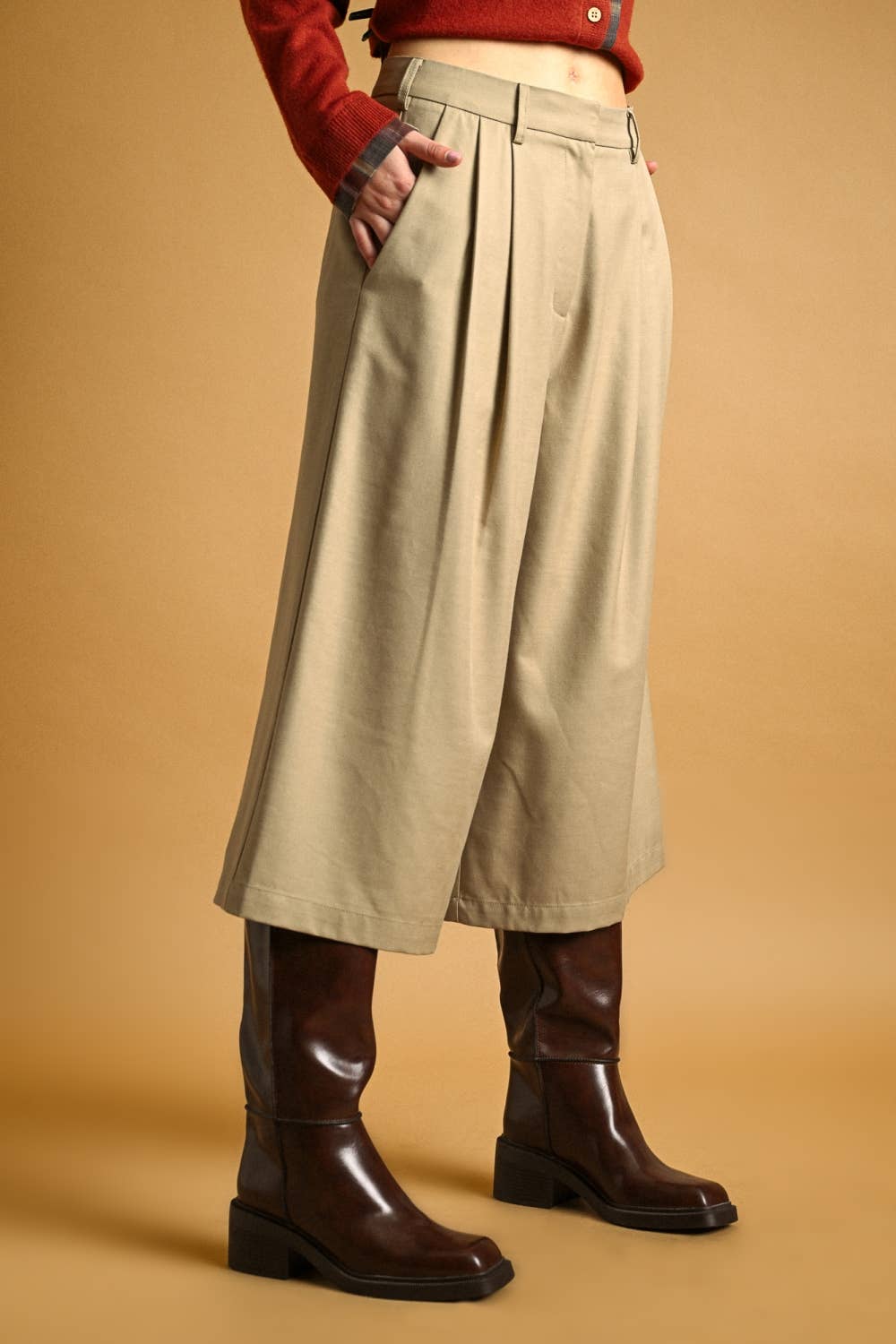 A model is shown from the waist down wearing high-waisted, wide-leg pleated cropped trousers in a light khaki-beige color, styled with a red cardigan and dark brown leather boots against a solid warm tan background.