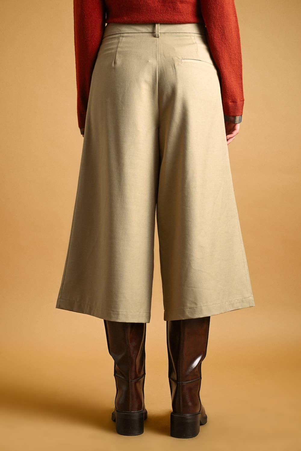 A person shown from the back wearing high-waisted, wide-leg cropped pants in a light beige/khaki color, paired with a rust-red sweater and dark brown knee-high boots, against a solid warm tan background.