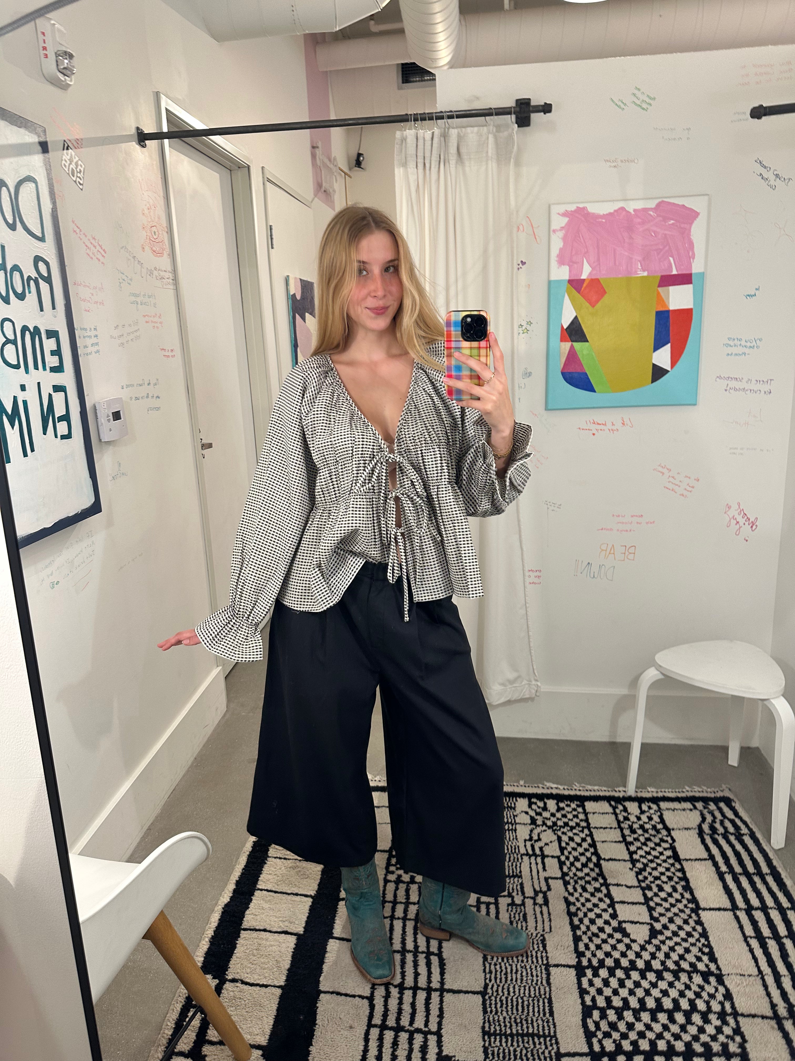 A woman takes a mirror selfie in a room, wearing a gray and white vertically striped long-sleeve blouse with a tie neckline, black wide-leg pants, and teal boots.