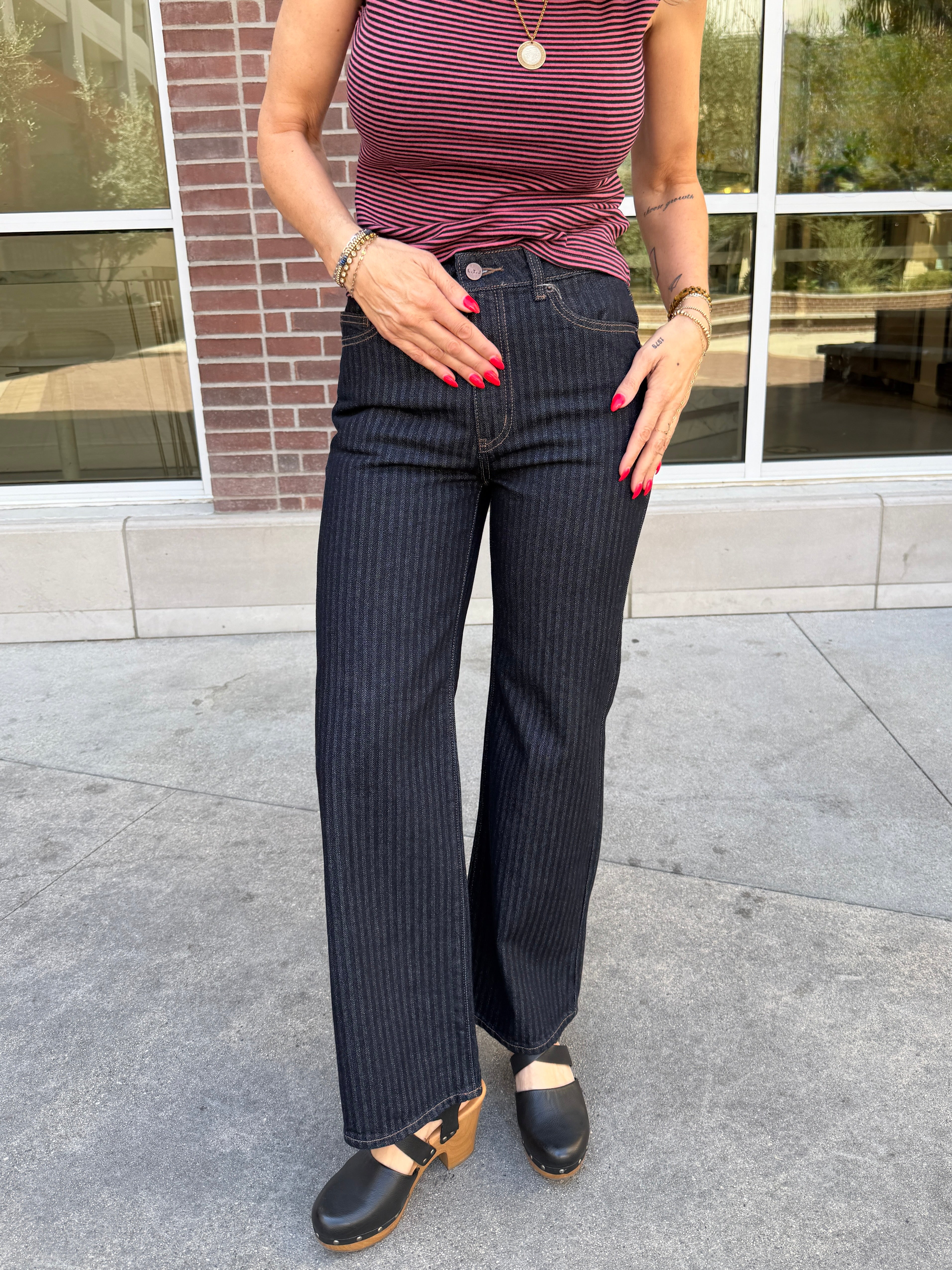 A person wearing dark navy pinstriped jeans, a burgundy and pink striped top, and black clogs, standing outdoors in front of a brick building with large windows.