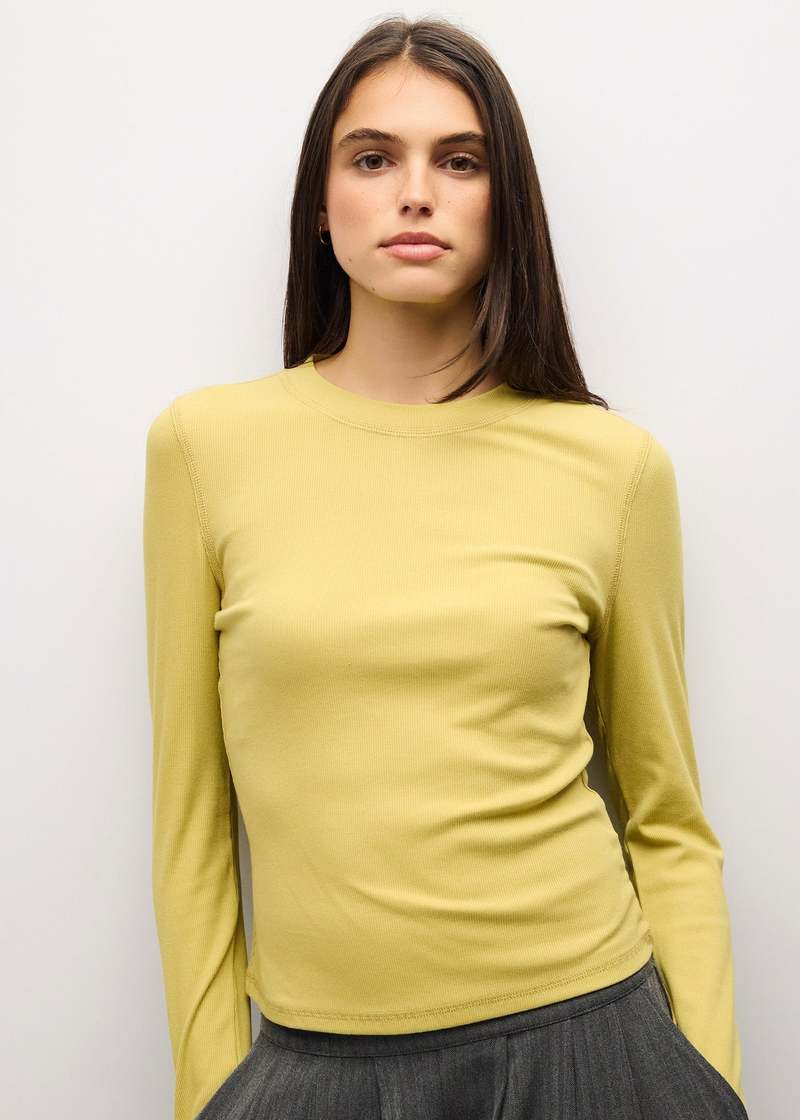 A woman wearing a fitted, long-sleeve crewneck top in a soft pastel yellow, paired with a dark gray skirt, standing against a plain light gray background.