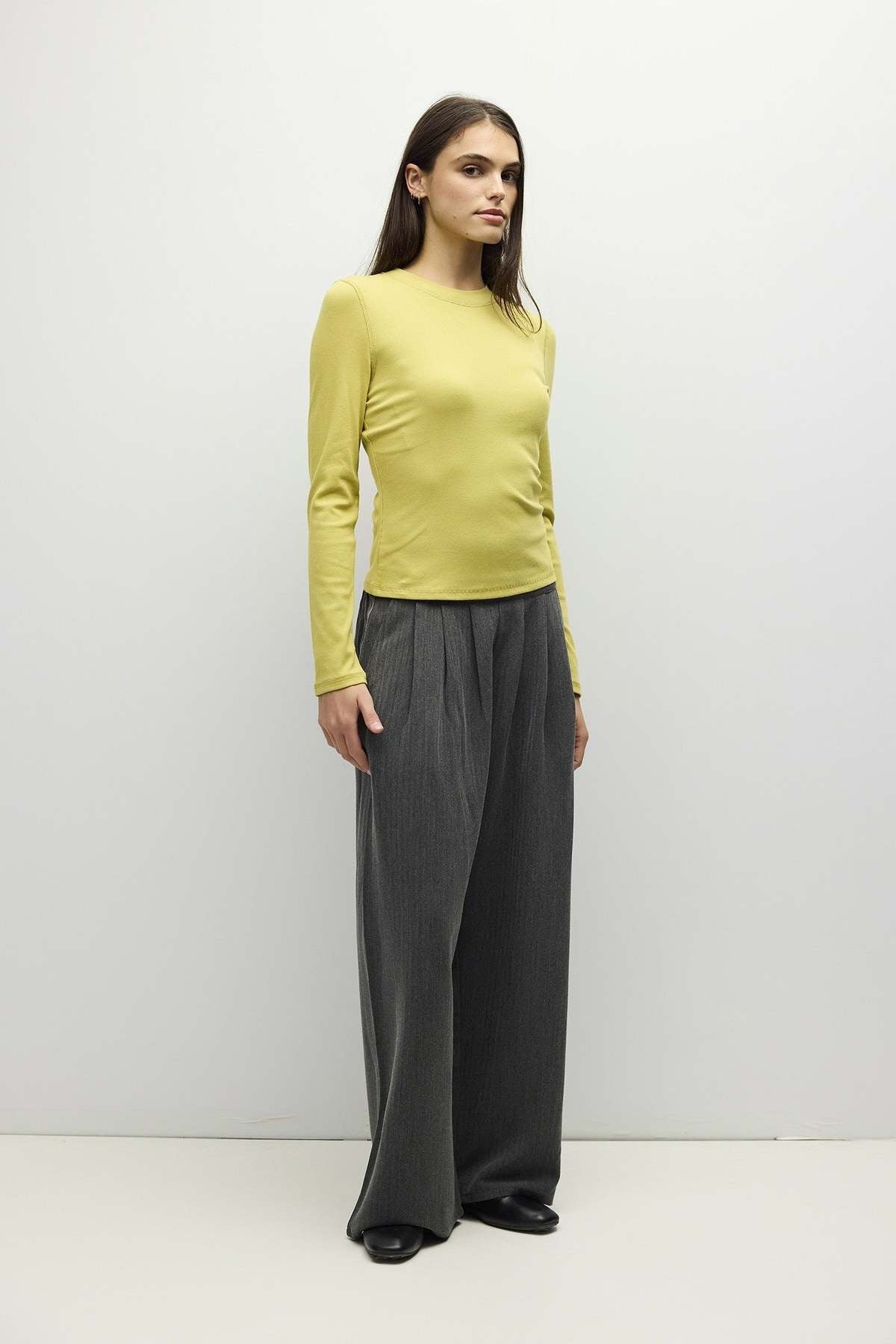 A woman modeling a fitted long-sleeve top in pale yellow-green paired with wide-leg dark gray trousers, standing against a plain light gray studio background.