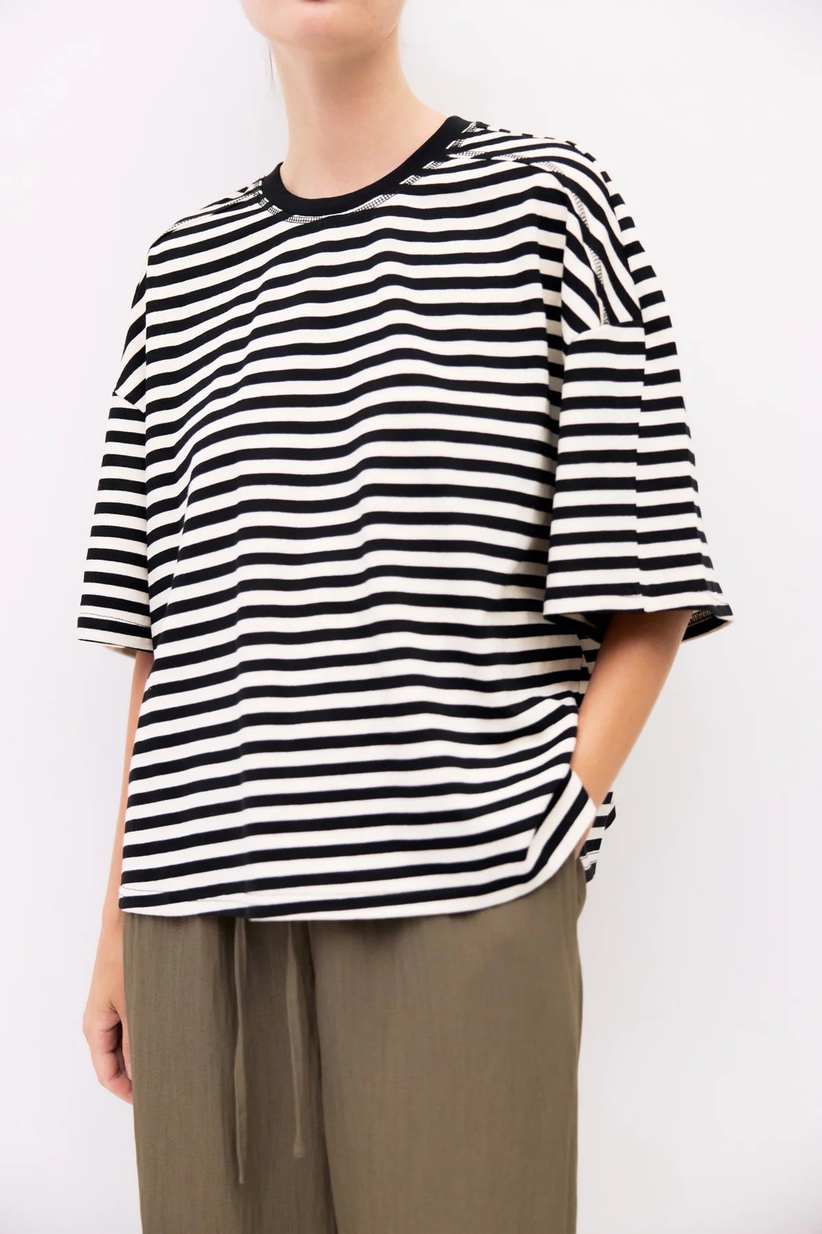 A person wearing an oversized black and white horizontally striped short-sleeve top with a black crew neck, paired with olive green drawstring pants, against a plain white background.
