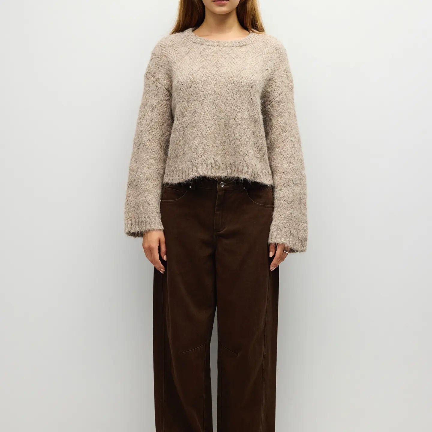 A woman wearing a fuzzy cropped knit sweater in a light taupe-beige color, paired with dark brown wide-leg pants, standing against a plain light gray background.