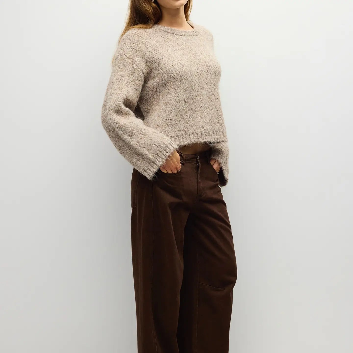 A woman wearing a chunky, fuzzy knit sweater in a light oatmeal beige color and dark brown wide-leg trousers, standing against a plain light gray wall.