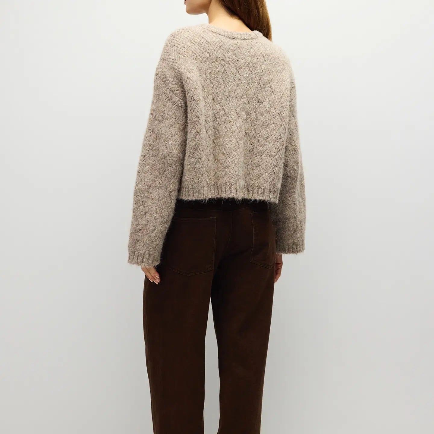 A woman shown from the back wearing a cropped, textured beige knit sweater and dark brown pants against a plain light gray background.