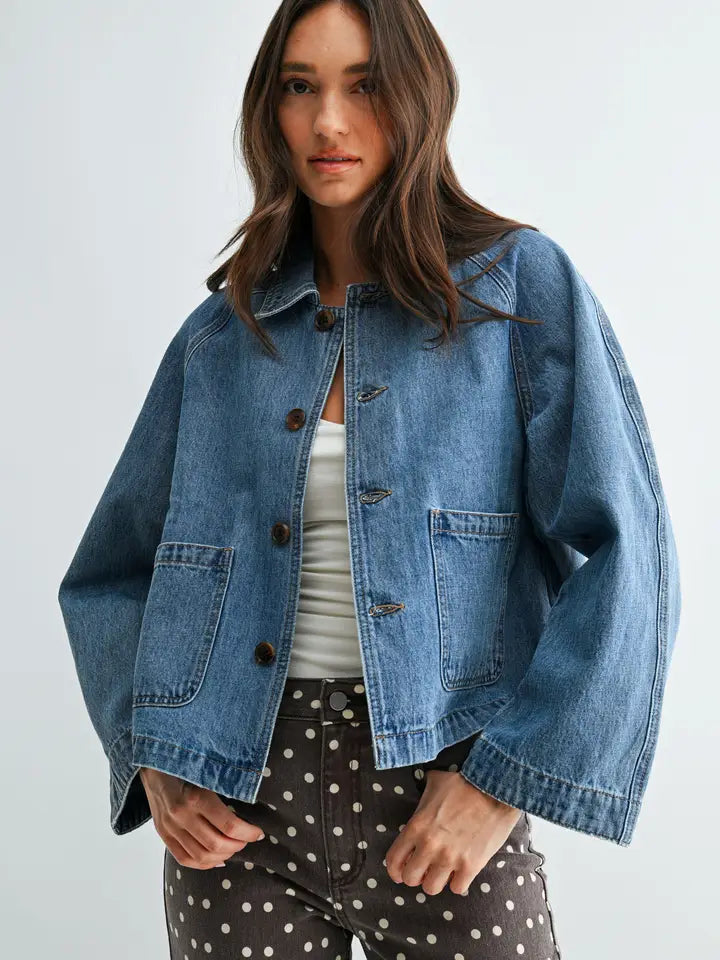 A woman wearing a cropped, oversized light blue denim jacket over a white top and dark brown polka-dot pants, photographed against a plain light gray studio background.