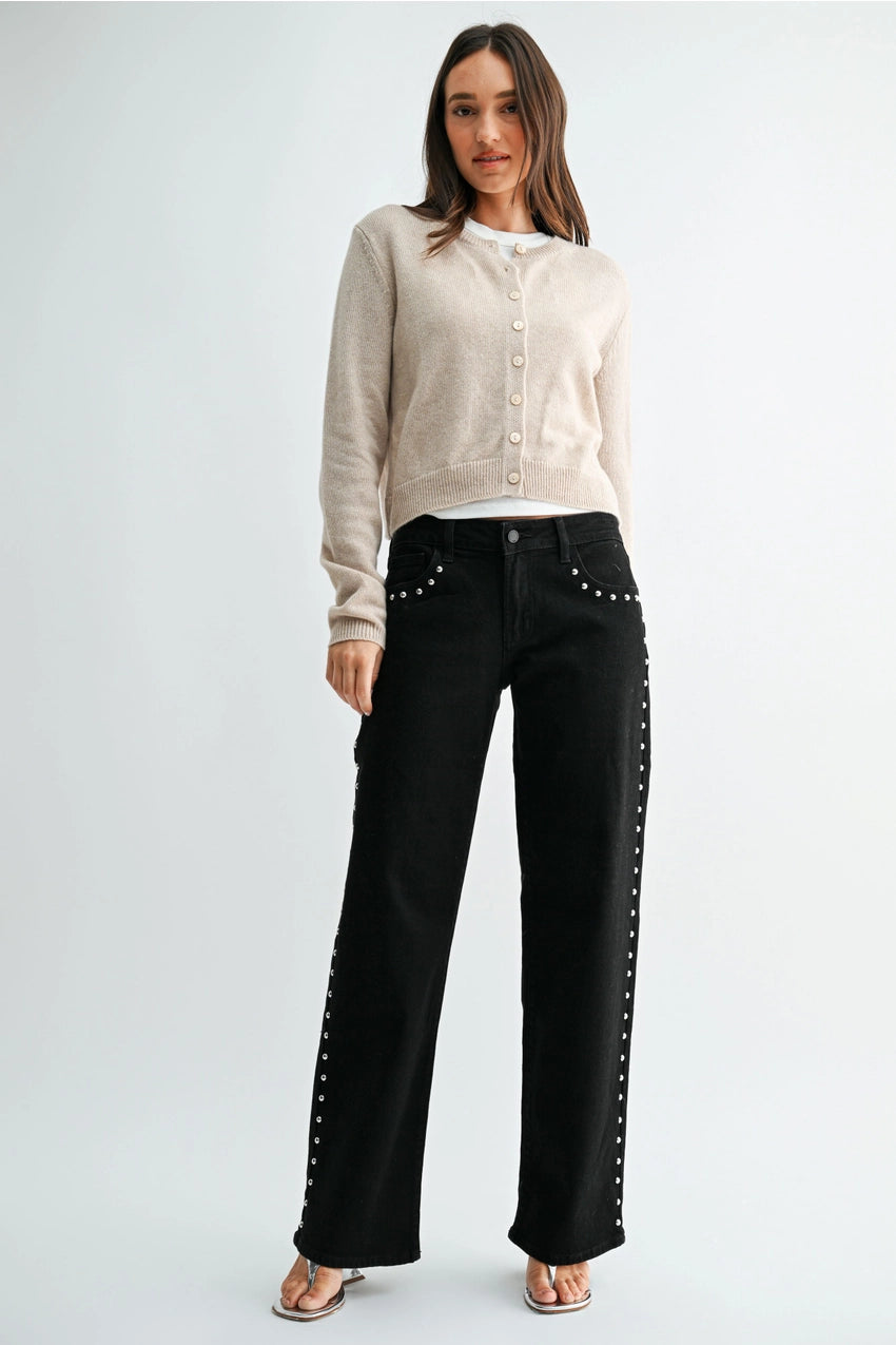 woman wearing black jeans with silver studs and a cream sweater in front of a white background. 